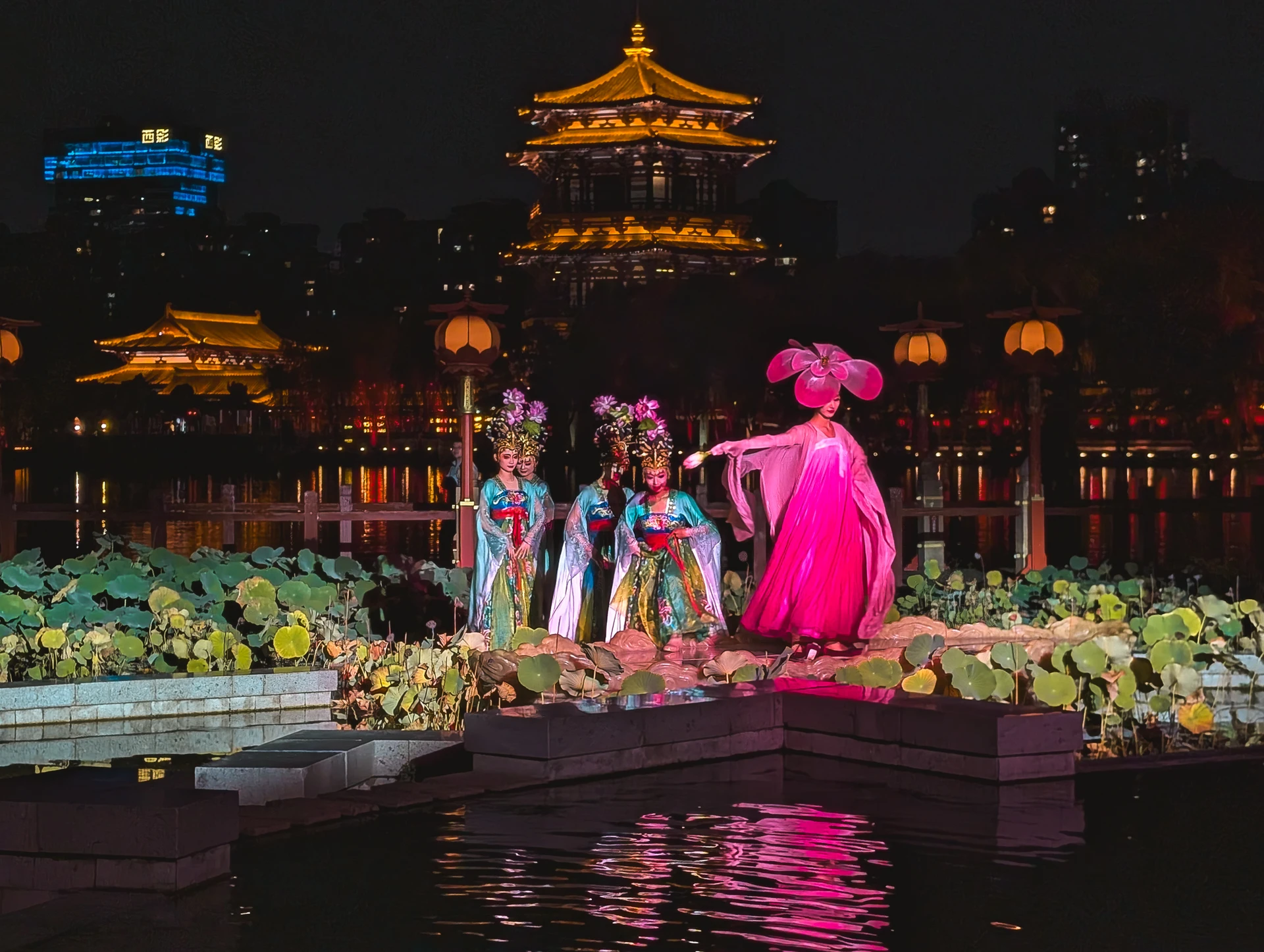 Performers in colourful costumes on a lakeside stage beneath an illuminated tower