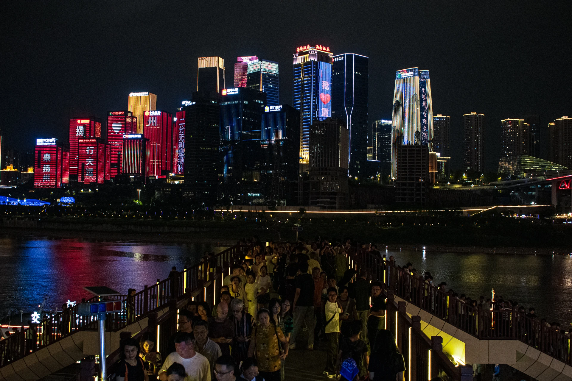 Crowds on a waterfront promenade facing colourful illuminated skyscrapers