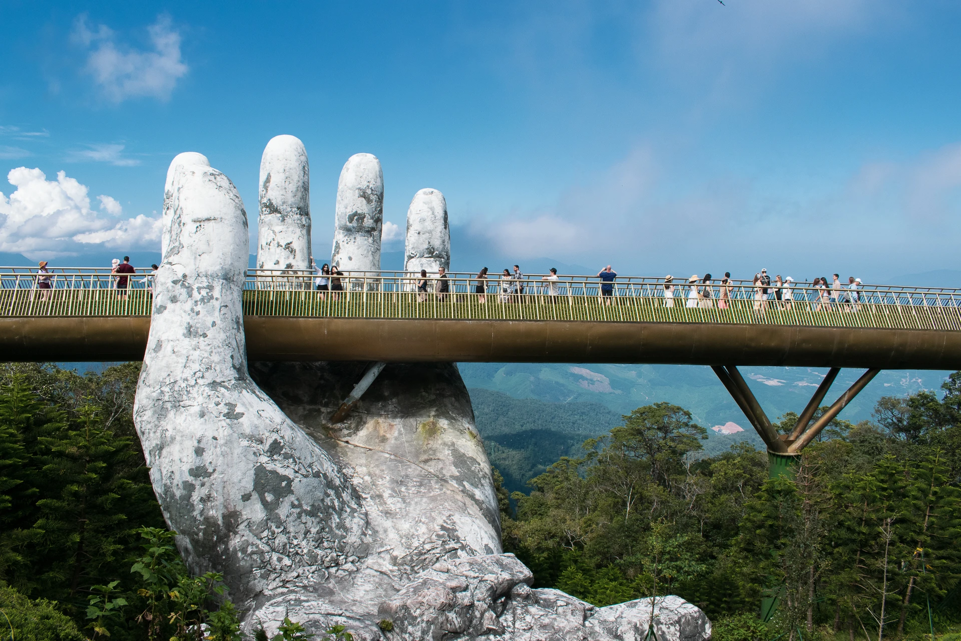 Visitors crossing a golden footbridge held by giant stone hands above forested hills