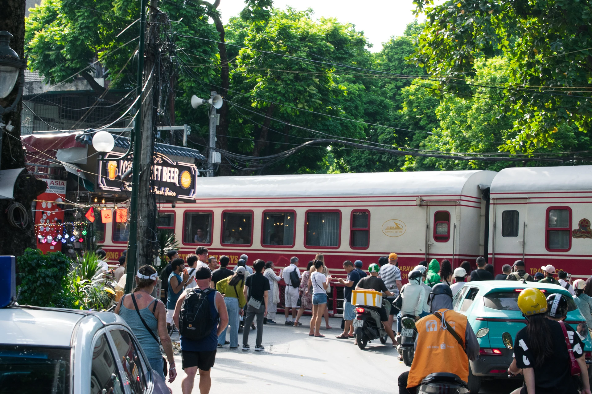Pedestrians and motorbike riders waiting as a red-and-cream train crosses a busy street